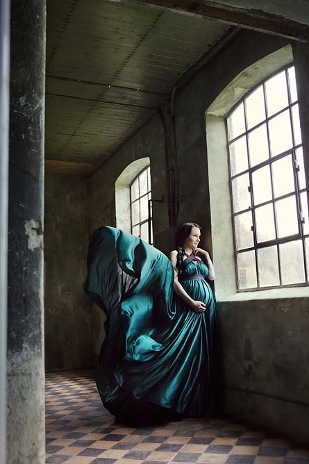 Elegant gravidfoto av en kvinne i blå kjole i industribygg med store vinduer, som fremhever mor- og barneglede.