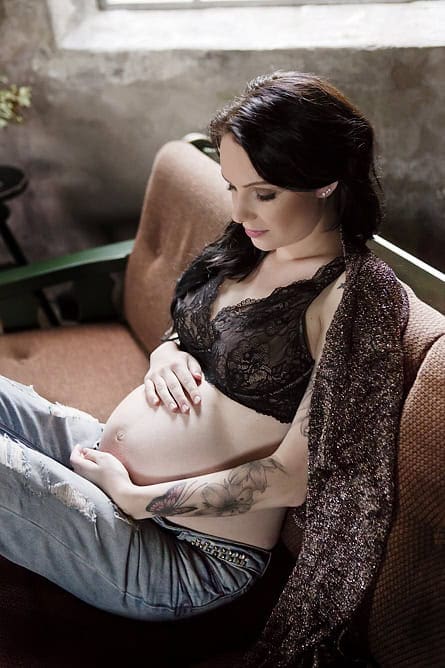 Lysbildende gravidfoto av en gravid kvinne som sitter på en sofa, iført svartlingende undertøy og skinnende sjal, som viser sin gravide mage i en rolig og intim setting, perfekt for gravidfotografering.