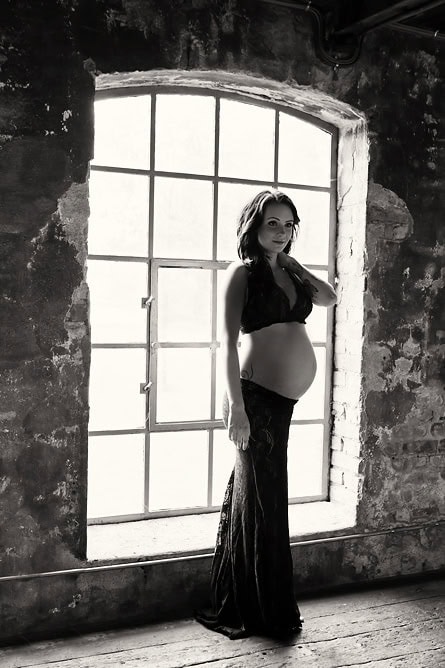 Elegant gravidfotografering av gravid kvinne foran stor vindusramme i industrielt preg, fokus på magens skjønnhet og modenhet. Perfekt for gravidebilder som fremhever graviditetens magi.