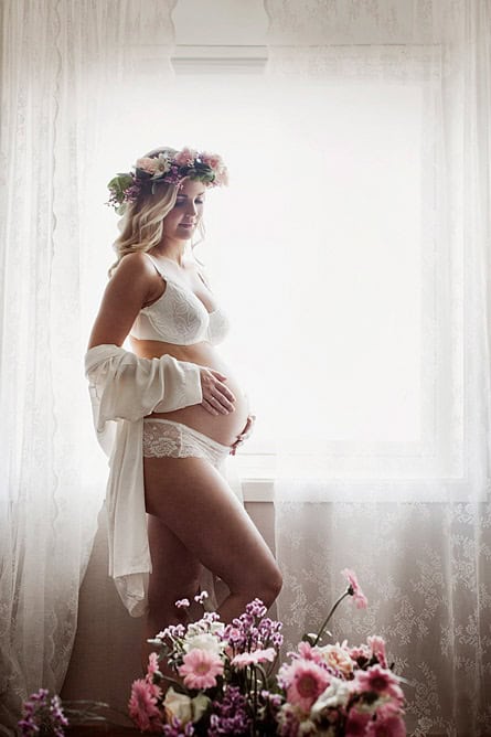 Kvinne i gravidfoto med blomsterkrans står ved et vindu i mykt lys, ikledd hvile klær, omgitt av blomster, foran et lyst gardin.