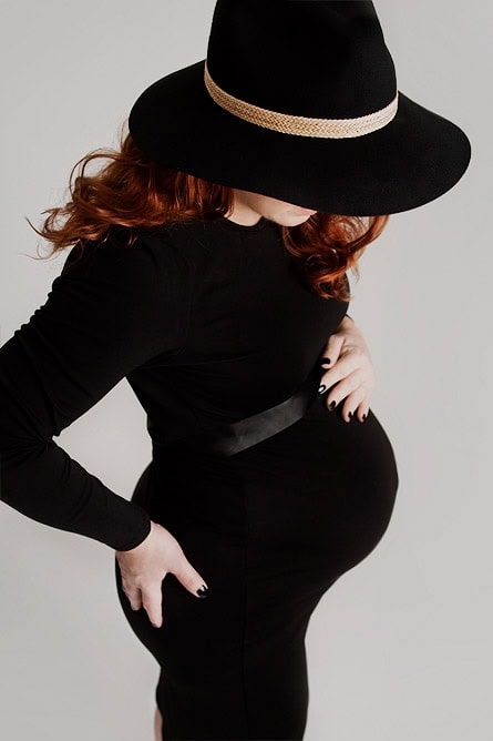 Elegant gravidfotografi av en gravid kvinne i svart kjole og bredbrun hatt, fremhever den vakre formen til fosteret med mykt lys.