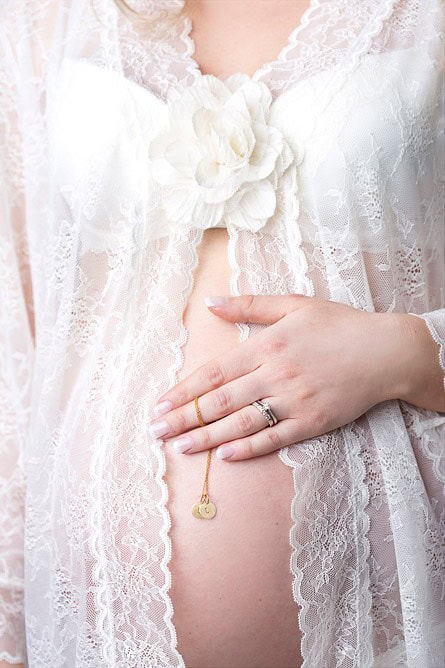 Nydelig gravidfoto av kvinne i hvit blonderomantisk kjole med hånd på mage og graviditets smykke, perfekt for gravide fotosesjoner og minneverdige øyeblikk.