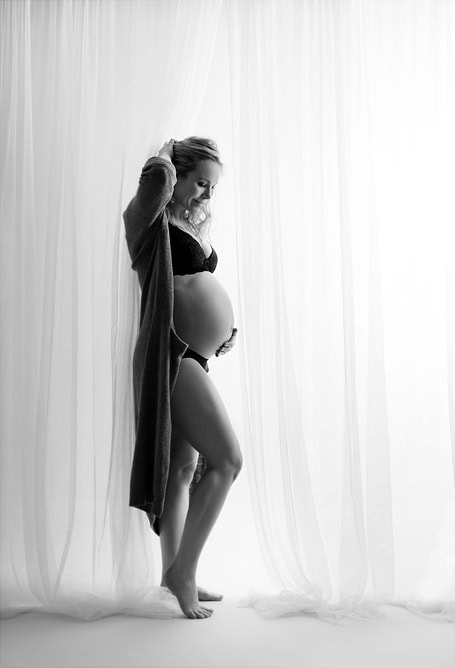 Elegant gravidfoto av gravid kvinne i svart og hvitt, opplyst av mykt dagslys fra bakgrunnslyset, som fremhever den gravide magen, perfekt for gravidfotos og minneverdige øyeblikk.