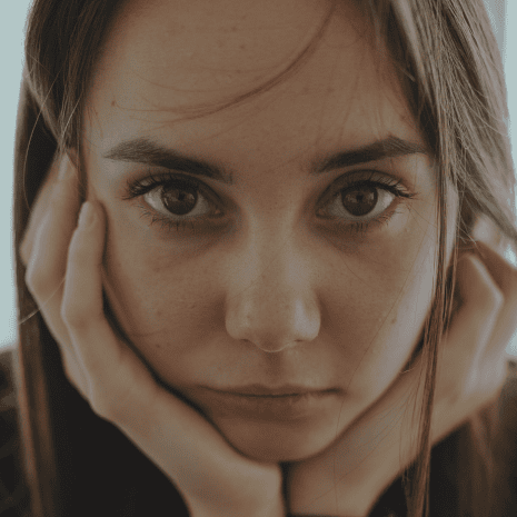 woman with long brunette hair holding her head in her hands as she stares into the camera.