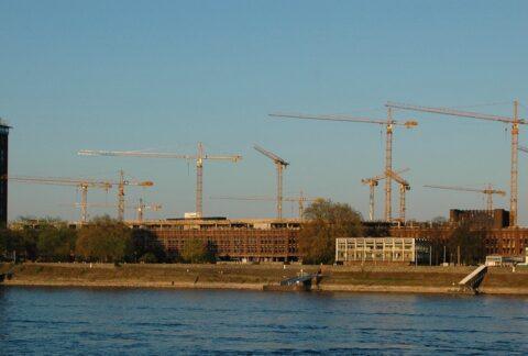 Bauprojekt mit mehreren Kränen und einem historischen Gebäude am Flussufer.