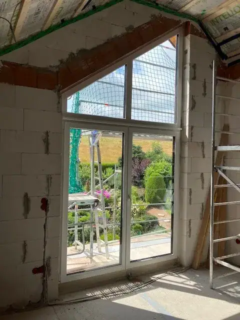 Weiße Balkontür mit integrierten dreieckigen Oberlichtern von Fütterer Fenster & Türen GmbH, in einem Rohbau installiert, bietet einen weiten Blick in einen grünen Garten.