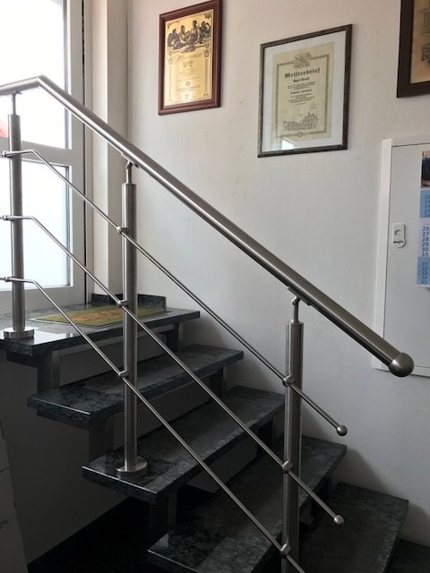 Modernes Treppengeländer aus gebürstetem Edelstahl von Stahl- und Metallbau Drost mit vertikalen Pfosten und horizontalen Stäben, montiert an einer Treppe mit dunkelgrauen Granitstufen.