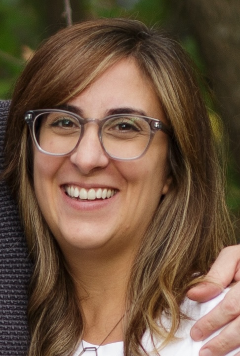 Smiling woman with shoulder-length light brown hair and clear-framed glasses outdoors.