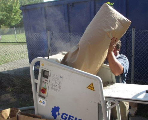 Person kippt einen Sack mit losem Dämmmaterial in eine Einblasmaschine im Freien; dabei staubt das Material