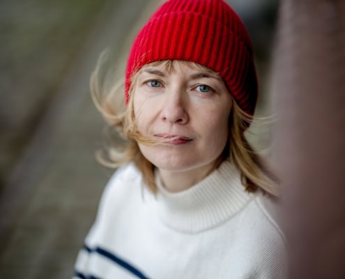 Authentisches Frauenporträt an einem windigen Tag mit einer roten Mütze als Farbakzent. Die Aufnahme fängt einen nachdenklichen Moment in urbaner Umgebung ein.