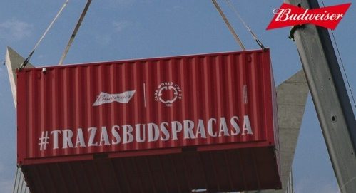 Imagem mostra o icônico contêiner vermelho da Budweiser com a hashtag #TRAZASBUDSPRACASA sendo içado por um guindaste, marcando o início da ação promocional em São Paulo e no Rio de Janeiro. A campanha oferece brindes exclusivos sem necessidade de compra, promovendo a experiência da marca diretamente nas ruas das capitais.