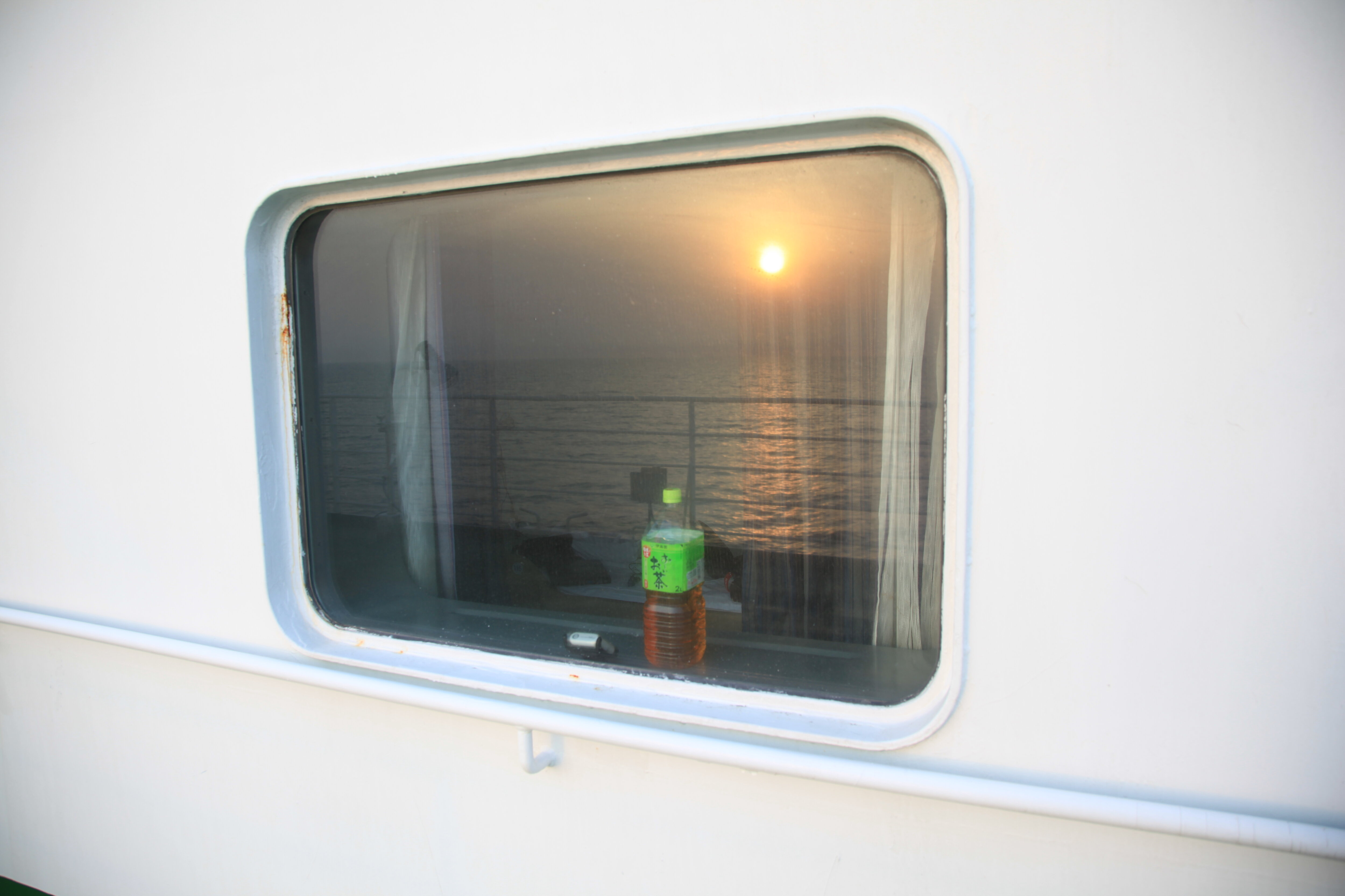 船室窓越しに朝日とお茶のペットボトル