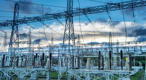 An outdoor electrical substation with multiple power lines, metal towers, and transformers set against a cloudy sky at sunset. 