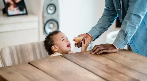 Toddler opens mouth crying, reaching toward the cup held by an adult