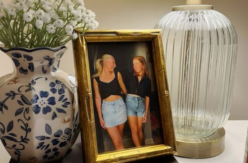 Gold‑toned nightstand styled with the book ‘Sweet Caroline,’ a blue and white vase, gold photo frame of two friends, and a lamp — chic and personal bedside table decor ideas from By Paige Nicole.