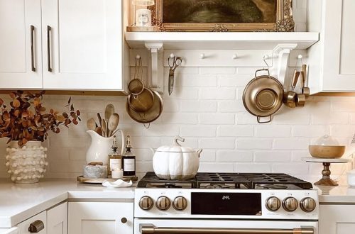 White kitchen and stove decorated with brass pots, pans, and a pumpkin kettle — cozy fall‑inspired top of stove decor ideas from By Paige Nicole.