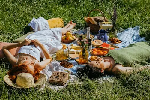 Wohlbefinden durch Achtsamkeit und Naturgenuss Zwei Frauen liegen entspannt auf einer Picknickdecke in einer hohen Sommerwiese neben einem Korb mit frischem Obst und Gebäck; finden Sie Ihre innere Balance mit Vitawy durch Achtsamkeit, Entspannung und Strategien für mentale Stärke.