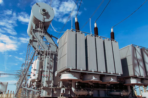 Large electrical transformer with cooling fins and high-voltage insulators under a blue sky, surrounded by power lines at an outdoor electrical substation.