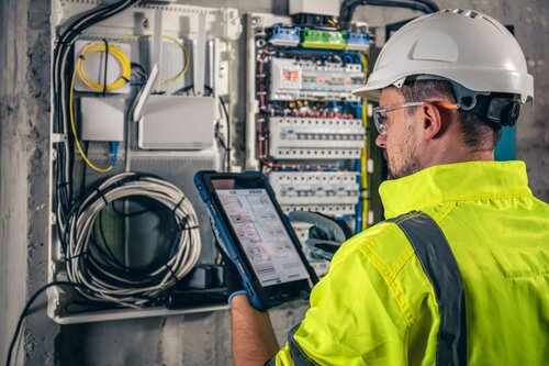 An electrician in a high-visibility jacket and hard hat uses a tablet to inspect an open electrical panel with wires and components on a wall.