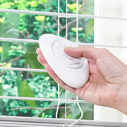 Hand holding remote control near window sensor on white window grill.