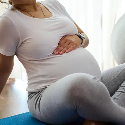 Holistic Pragnancy Care-A close up, cropped shot of a pregnant woman wearing a white t-shirt and grey leggings, sitting on a blue yoga mat. She has one hand resting gently on her stomach, and a white exercise ball is visible in the background.