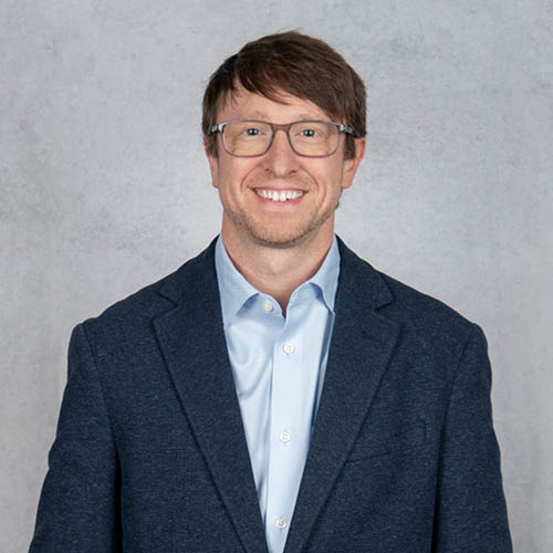 Professional headshot of David Rosenberger wearing glasses, a navy blazer, and a light blue dress shirt against a neutral gray background.