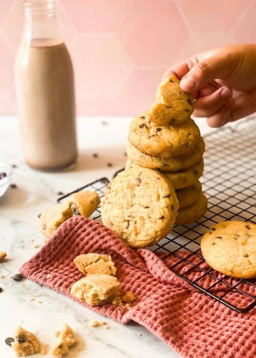 stack of SUGAR COOKIE WITH CHOCOLATE CHIPS on a wiring rack and pink towel, a hand holding one cookie, around a bottle of chocolate milk and chocolate chips