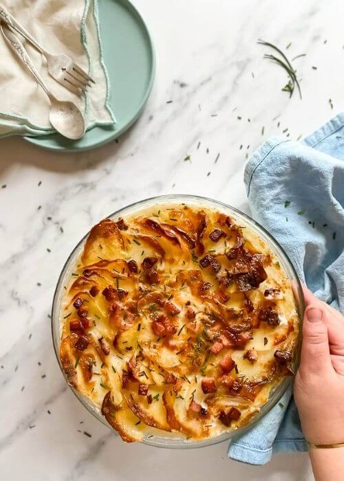 air fryer scalloped potatoes in a tray, under a blue towel, on the side a plate with a towel and cutlery, under a marble background, hand holding the tray