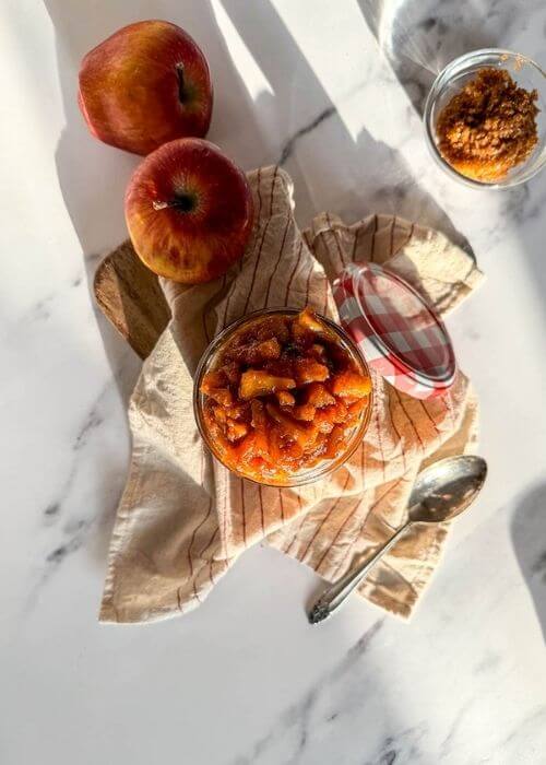 Delicious Apples Caramelized With Cinnamon And Brown Sugar, Vegan & Easy 5 apples caramelized in a jar, over a striped towel, a spoon, apples and brown sugar on the sides, over a marble background