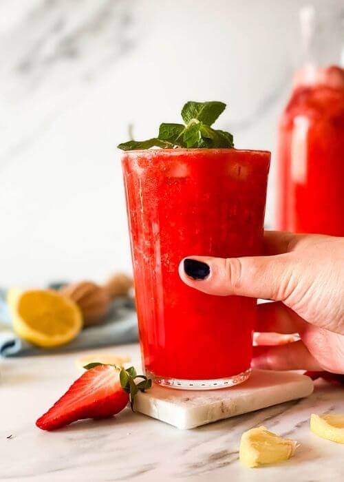 Easy Blended Strawberry Lemonade Recipe 4 hand holding glass of blended strawberry lemonade on a marble coaster, behind a jug of lemonade, strawberries mint and lemons around, in the back a blue towel and lemon squeezer on a marble background