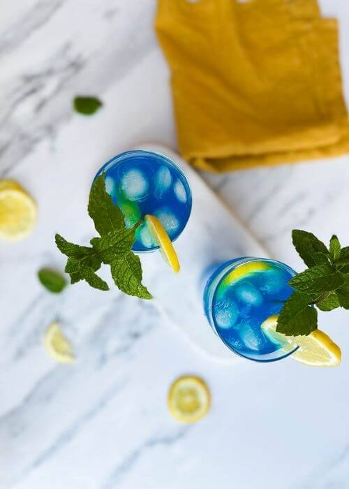 Summer Blue Lagoon Fizz Chillis Recipe Delicious Cocktail 3 from top - two glasses of blue lagoon fizz chillis recipe on a marble tray and marble background, around a yellow towel, a lemon and mint leaves