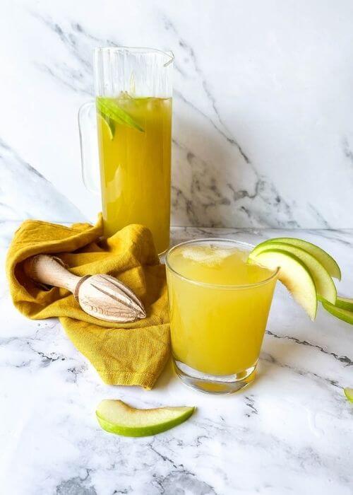 on a marble background, a glass of green apple mocktail, beside a yellow towel and lemon squeezer, behind a jug of green apple mocktail, slices of apples around