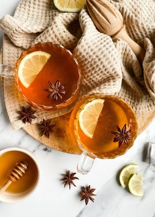 Festive Hot Toddy Mocktail Recipe, Warm & Delicious 6 from top two cups of hot toddy mocktail with slices of lemon and star anise, over a wooden board and light brown tea towel, slices of lemons, star anise and honey around,