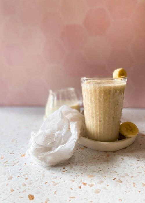 banana milk in a glass, on a tray with sliced banana, a towel, another glass and jug behind, a pink and speckled background