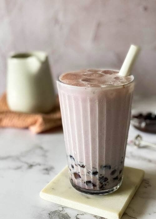 Taro Latte Recipe: A Quick & Delicious Summer Drink 1 taro latte in a glass with a straw, on a marble coaster, jug, towel and boba pearls in the background, on a marble background