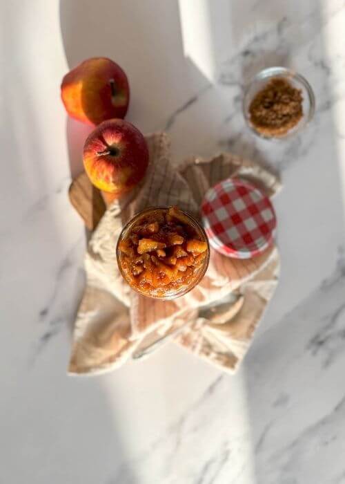 Delicious Apples Caramelized With Cinnamon And Brown Sugar, Vegan & Easy 2 from top - apples caramelized in a jar, over a striped towel, a spoon, apples and brown sugar on the sides, over a marble background
