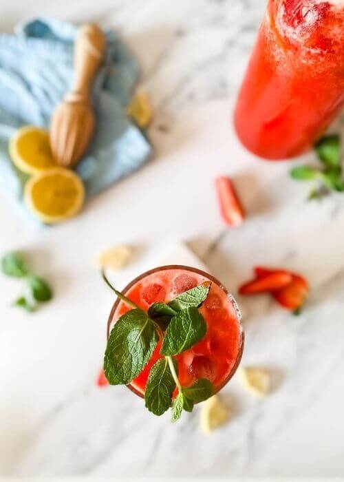 Easy Blended Strawberry Lemonade Recipe 3 from top - glass of blended strawberry lemonade on a marble coaster, behind a jug of lemonade, strawberries mint and lemons around, in the back a blue towel and lemon squeezer on a marble background