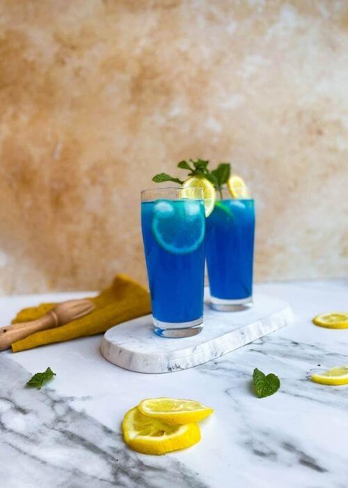Summer Blue Lagoon Fizz Chillis Recipe Delicious Cocktail 2 from front - two glasses of blue lagoon fizz chillis recipe on a marble tray and marble background, around a yellow towel, a lemon and mint leaves and lemon squeezer