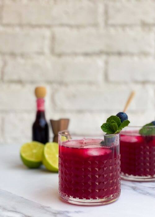 Amazing Blueberry Gin Cocktail, A 10-Minute Tasty Gin Tonic 4 two glasses of blueberry gin cocktail, in the back lime, blueberry syrup and measure cup, on a white and marble background
