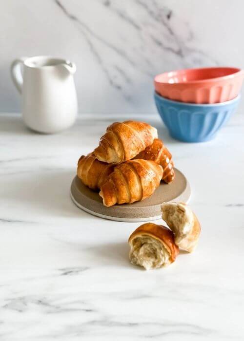 Croissant Air Fryer Recipe: Flaky, Delicious And Super Quick 2 A stack of shiny, air-fried croissants sits on a neutral-toned plate, with a broken croissant in front. In the background, a white milk jug and pastel-colored mixing bowls add a cozy kitchen vibe.