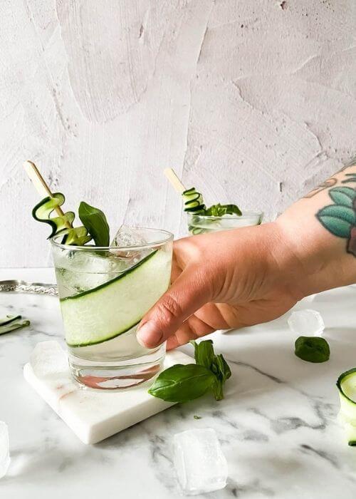 Easy Mocktail Recipes You Should Try 2 hand reaching for one two glasses of cucumber basil mocktail on marble coasters and on a marble background, around ice, basil, cucumber slices and an ice tongs