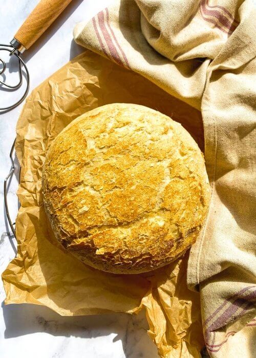 no knead bread, under parchment paper, wire rack, towel and on the side a bread a dough whisk, on marble background
