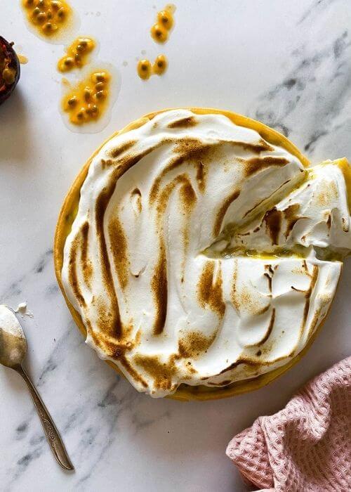 passion fruit meringue pie with a cut slice on a marble background, passion fruit and seeds on the side, spoon with meringue on the side