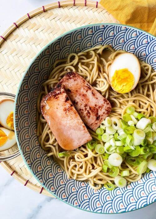 close up a bowl of ponyo ramen with noodles, spring onions, ham and egg. under a yellow towel, bowls with ingredients on a marble background