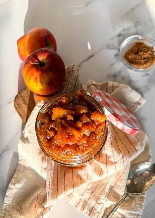 Delicious Apples Caramelized With Cinnamon And Brown Sugar, Vegan & Easy 6 close up apples caramelized in a jar, over a striped towel, a spoon, apples and brown sugar on the sides, over a marble background