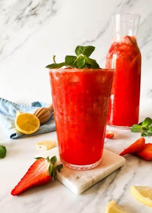 Easy Blended Strawberry Lemonade Recipe 1 glass of blended strawberry lemonade on a marble coaster, behind a jug of lemonade, strawberries mint and lemons around, in the back a blue towel and lemon squeezer on a marble background
