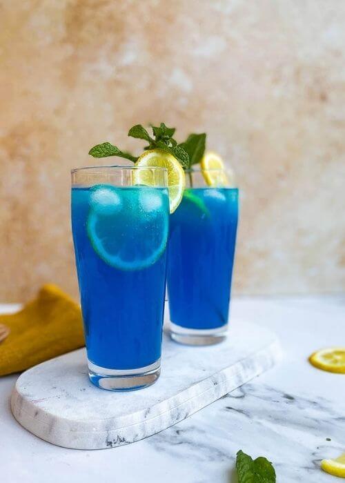 close up - two glasses of blue lagoon fizz chillis recipe on a marble tray and marble background, around a yellow towel, a lemon and mint leaves