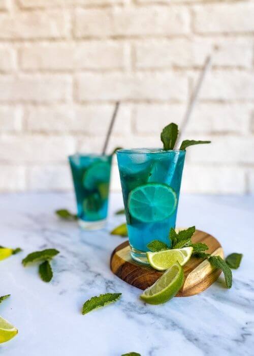 two glasses of blue mojitos on a wooden board, with lime slices and mint leaves on the side. On a white background