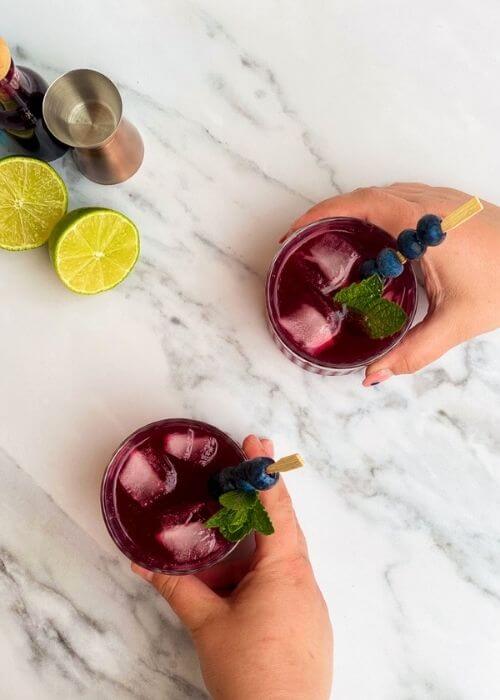 Amazing Blueberry Gin Cocktail, A 10-Minute Tasty Gin Tonic 3 from top, hands holding two glasses of blueberry gin cocktail, in the back lime, blueberry syrup and measure cup, on a white and marble background