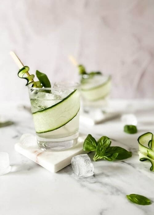 two glasses of cucumber basil mocktail on marble coasters and on a marble background, around ice, basil, cucumber slices and an ice tongs
